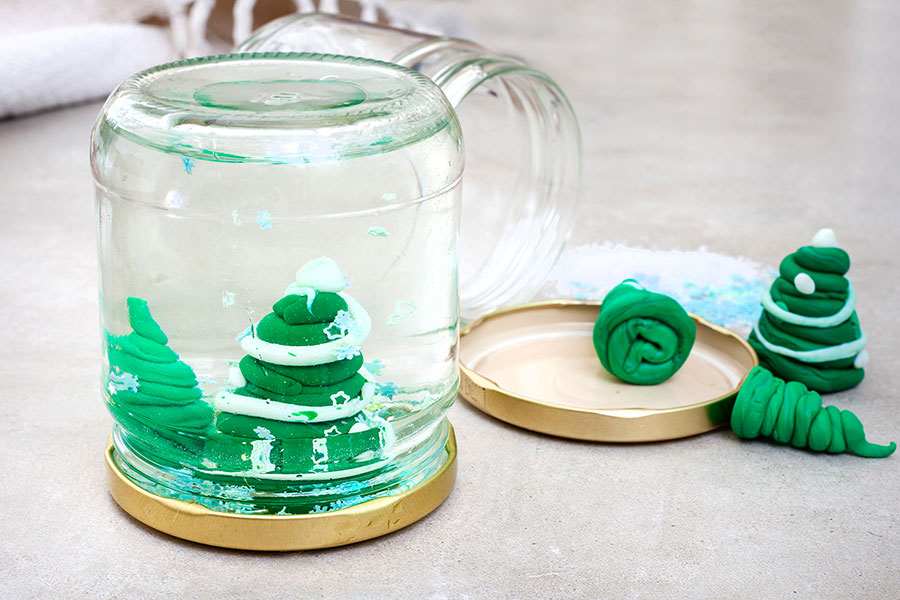 Das Bild zeigt ein DIY-Schneekugel-Projekt mit einem umgedrehten Glas, das auf seinem goldenen Deckel steht. Im Inneren des Glases befinden sich kleine grüne Weihnachtsbäume aus Knetmasse, Wasser und Glitzerpulver –  DIY-Ideen für Upcycling-Materialien